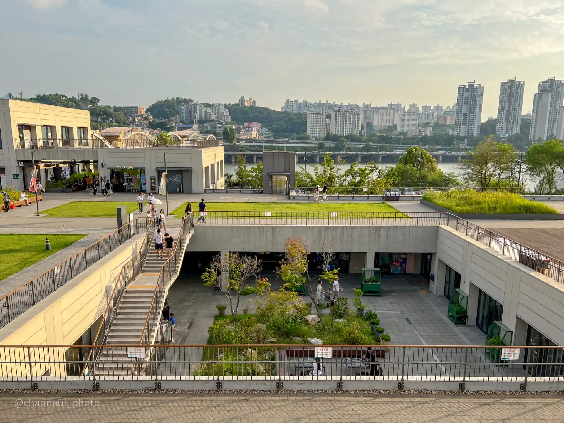 노들섬의 건물과 잔디밭, 사람들이 계단을 오르내리는 모습, 강과 고층 아파트가 배경에 보임.