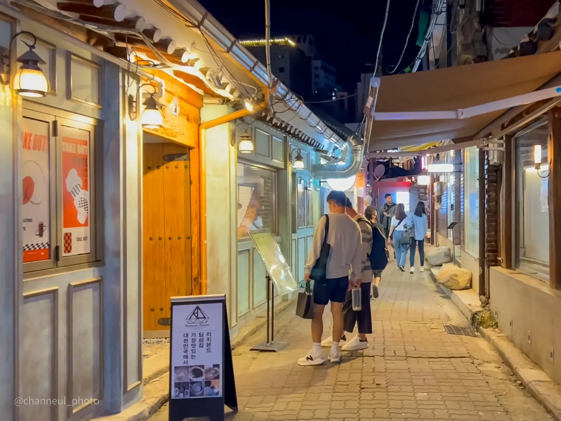 밤에 조명이 켜진 익선동 한옥 마을의 좁은 골목길에서 사람들이 걸어가고 있는 모습. 골목길 양옆으로는 한옥 스타일의 상점들이 늘어서 있고, 그 중 한 상점 앞에는 커피 테이크아웃 광고가 붙어 있다.