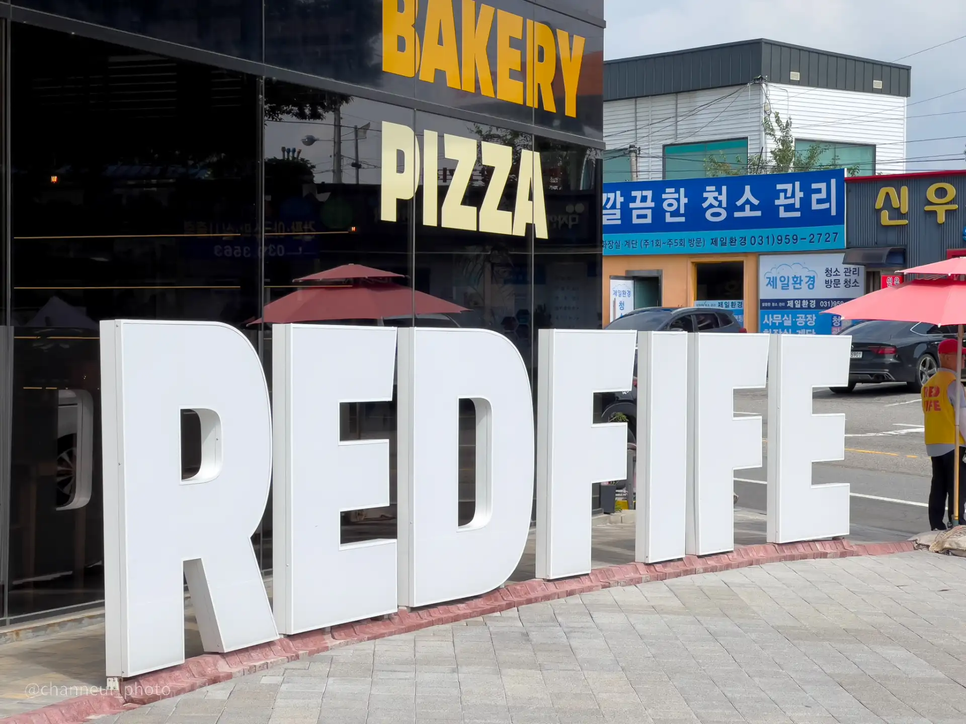 'REDFIFE'라는 큰 흰색 글자가 건물 앞에 설치된 모습이다. 건물 유리창에는 'BAKERY'와 'PIZZA'라는 노란색 글자가 보인다. 배경에는 상점과 가게들이 있으며, 빨간색 파라솔이 설치되어 있다.